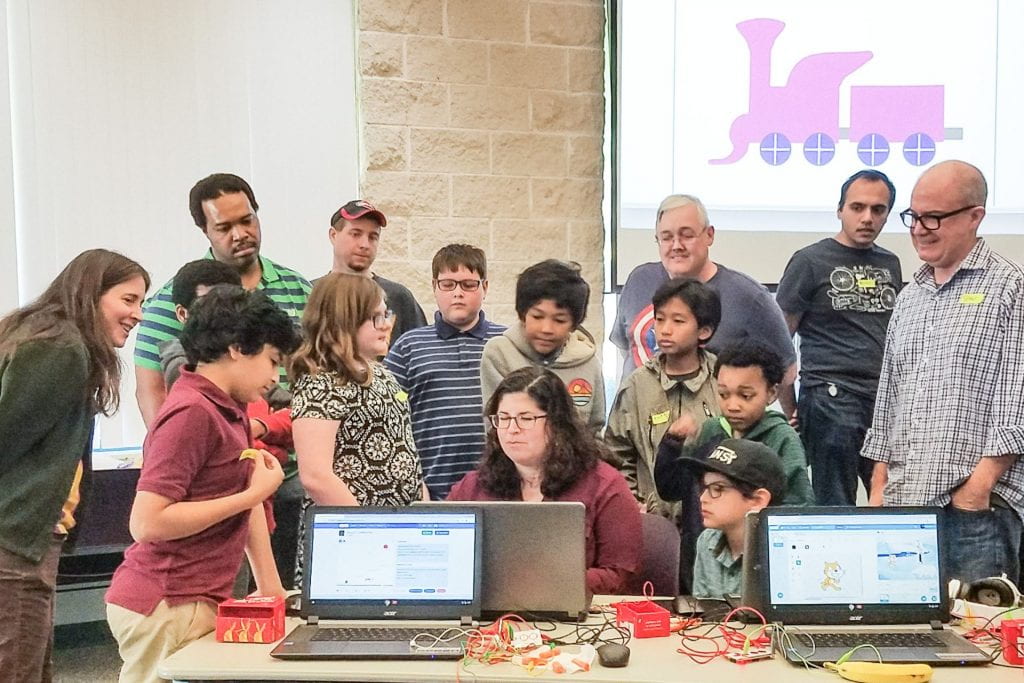 Adults and children gather around a laptop computer to watch a visual story.