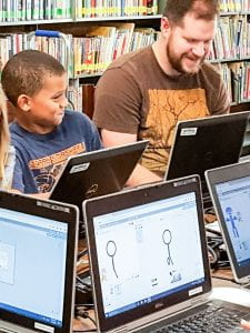 Father and son laugh while working together at a computer in a library.
